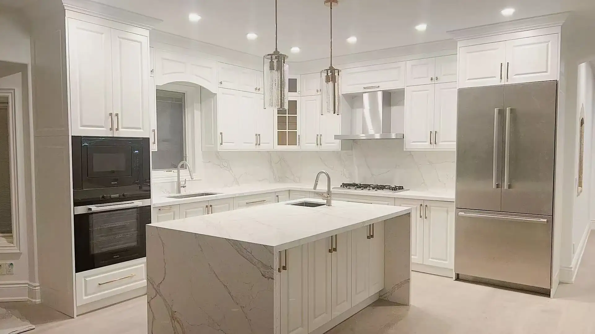 Marble-look waterfall island with a sink and a wide walkway between it and the fridge and wall ovens