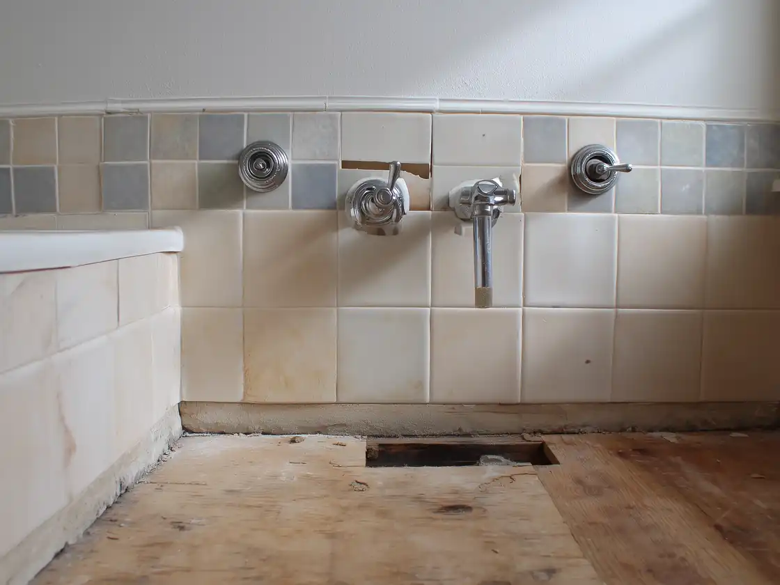 Bathroom wall and floor after vanity removal, with broken tile around the pipes and bare, damaged subfloor